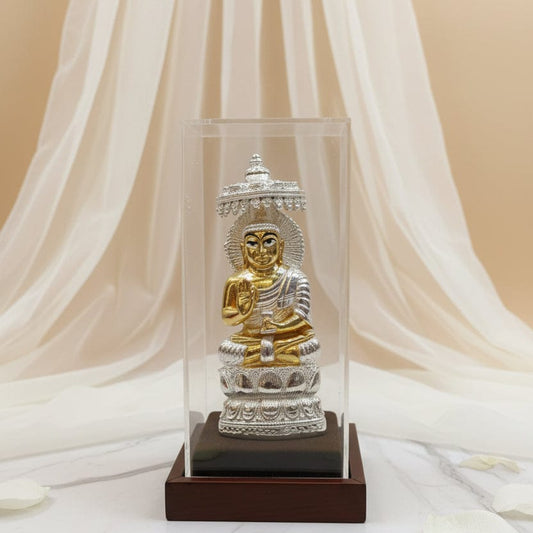 Silver and gold Buddha statue in a clear glass case with white fabric and petals in the background, featuring the brand 'KANUVEDA'.
